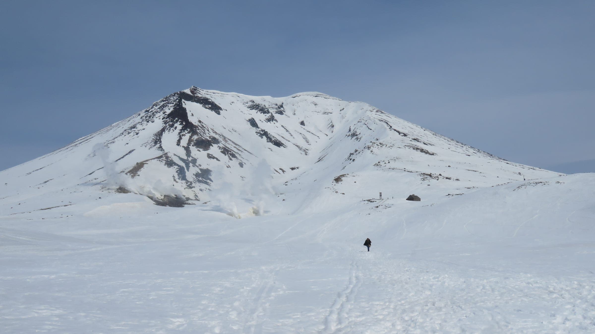 A lone skier climbs the snowfield toward Asahidake's summit cone under a pale spring sky