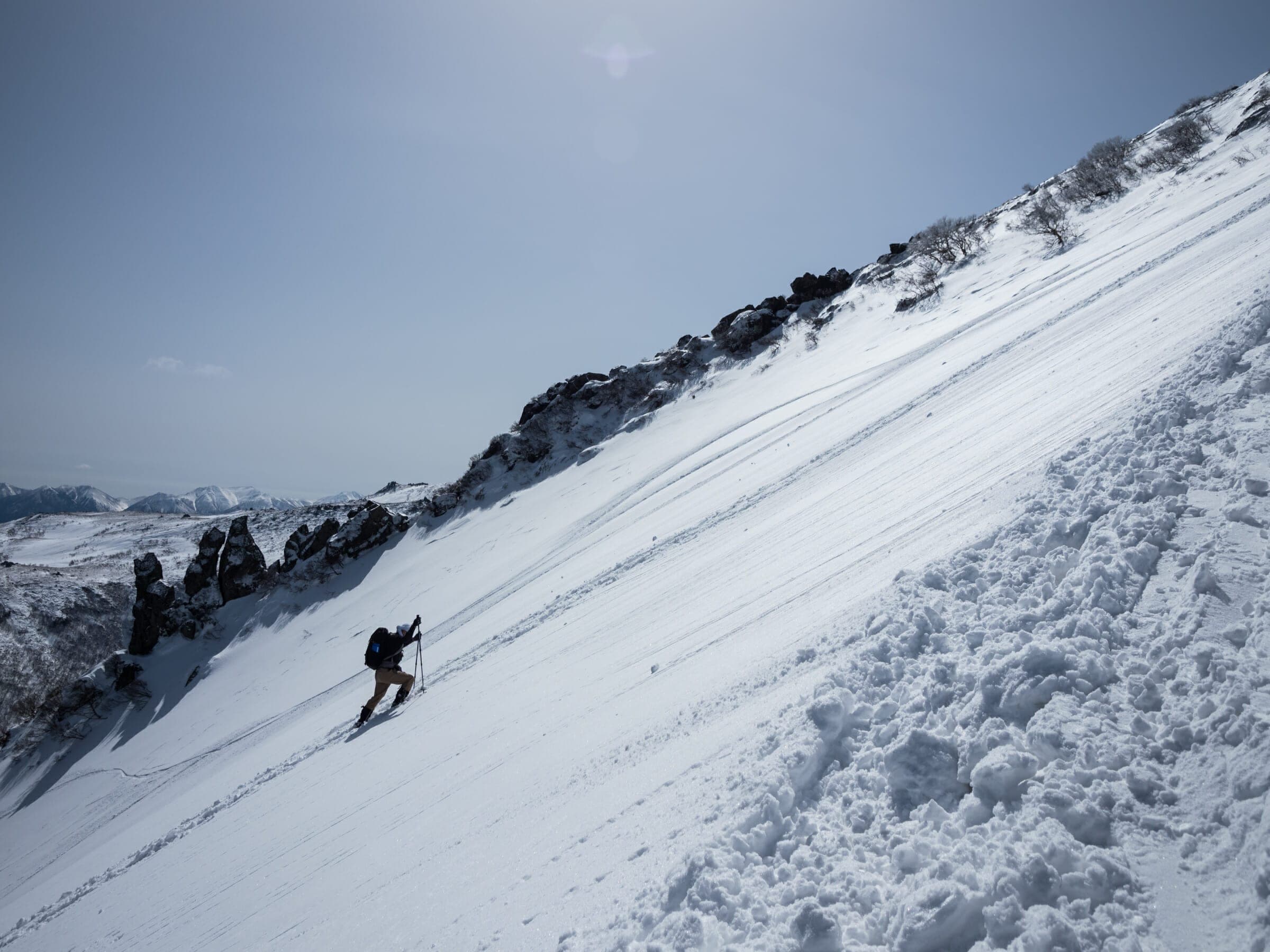 A skier descends a steep snow slope below the rocky summit of Kurodake
