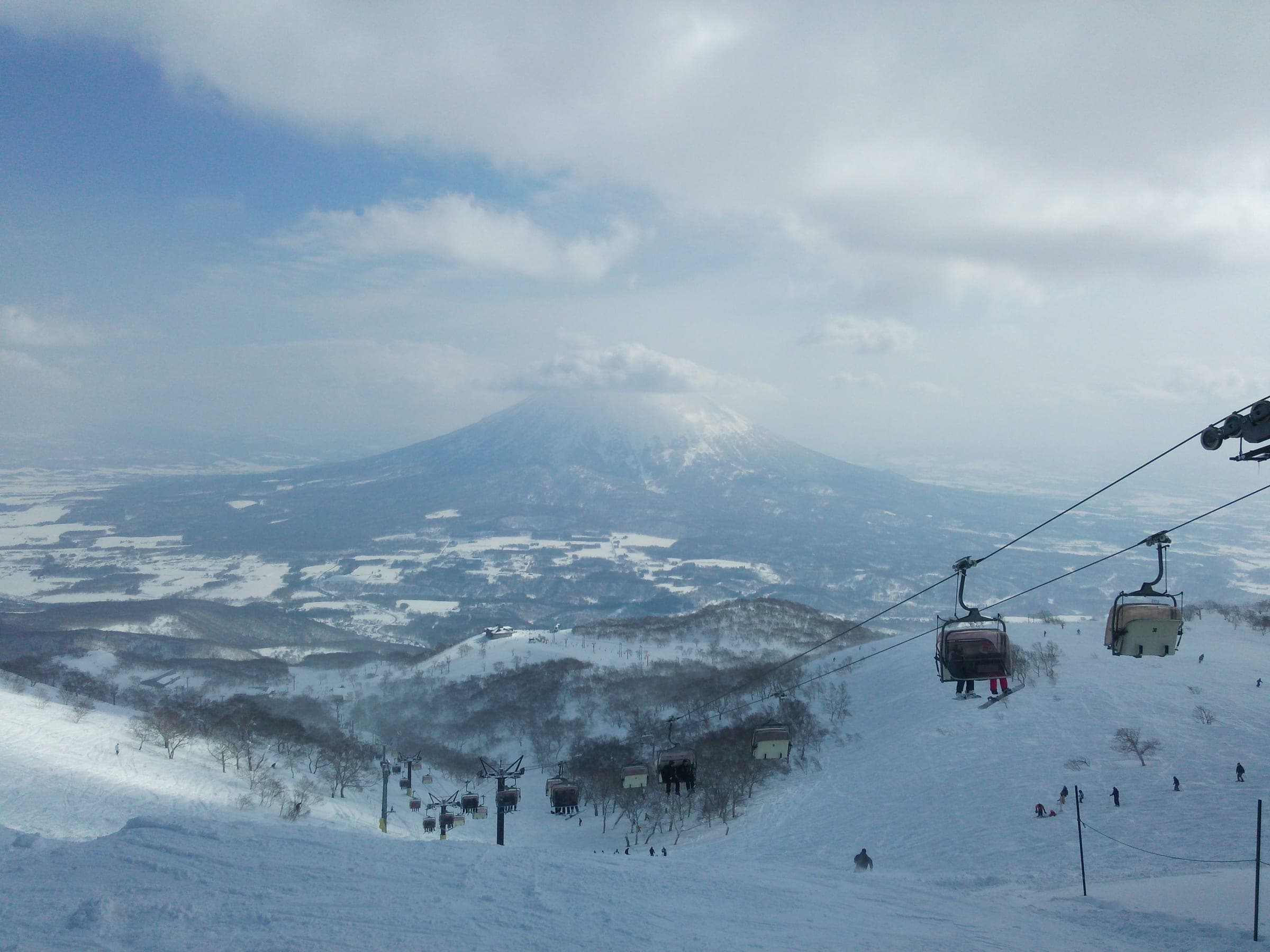 A hooded chairlift climbs Niseko Annupuri above snowy forest with Mt. Yotei in cloud