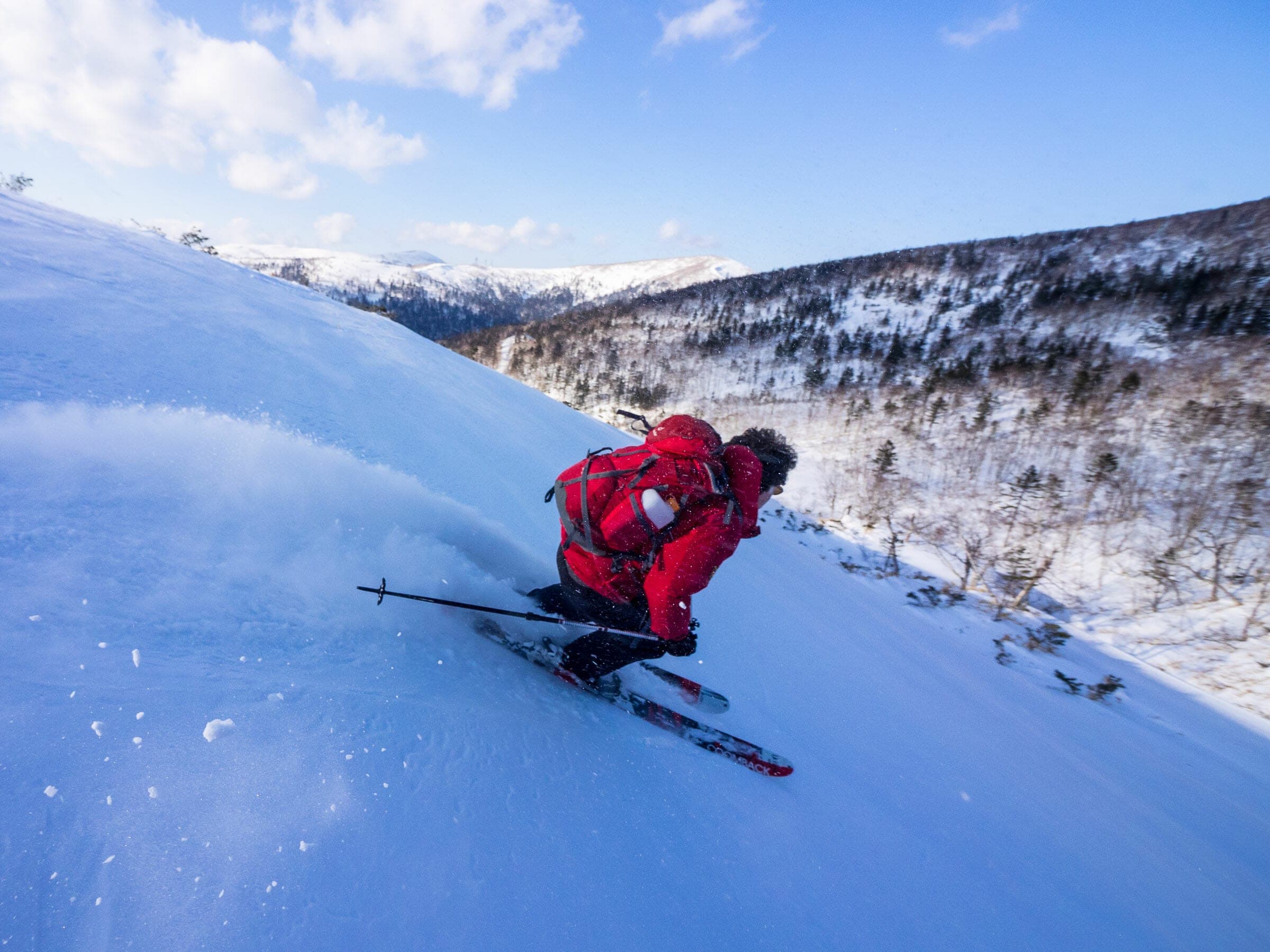A skier in red sprays snow on a steep sunny slope of Nissho Peak above Nissho Pass