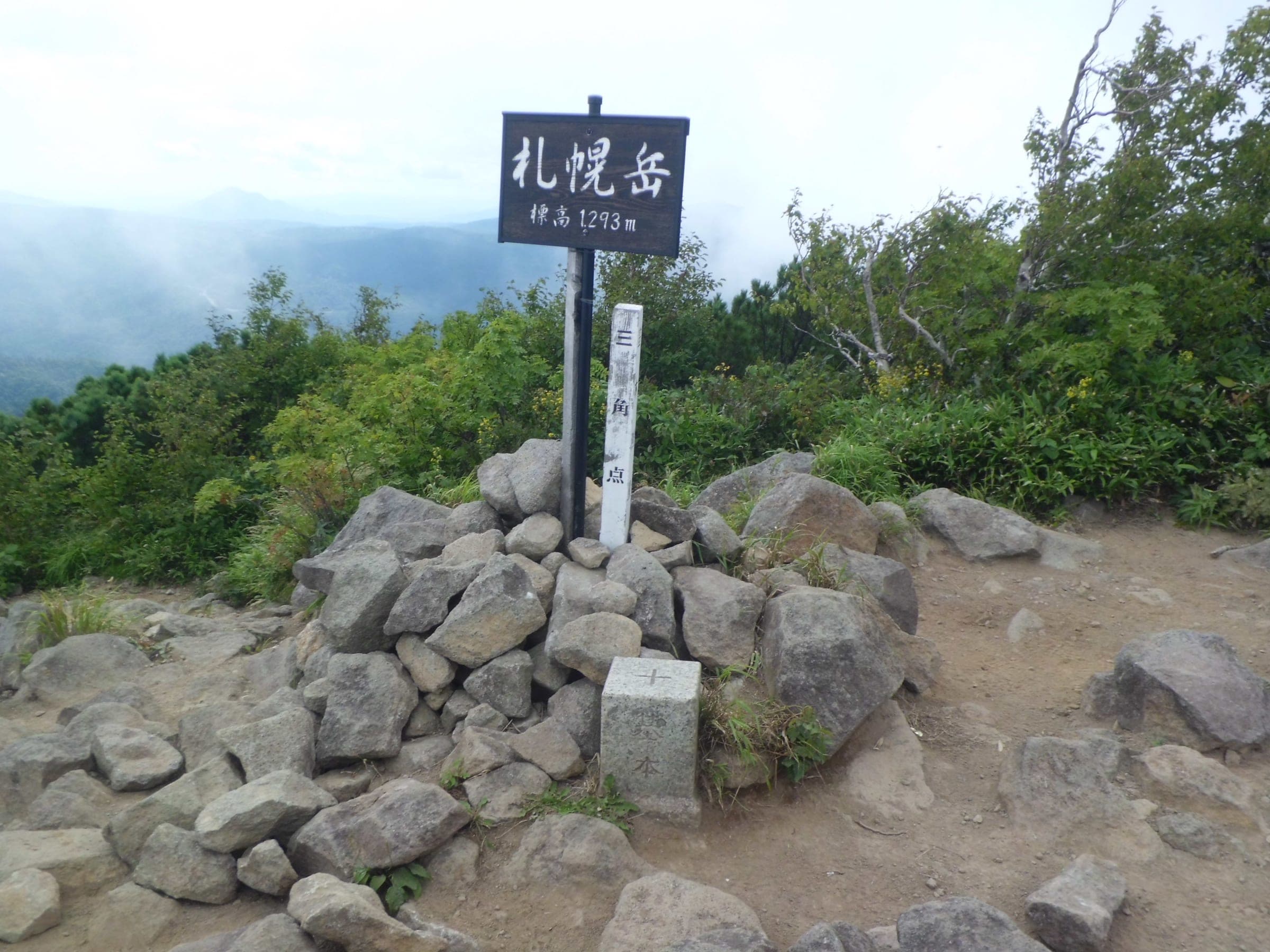 The summit marker of Sapporo-dake stands on a pile of rocks among summer scrub at 1,293 m