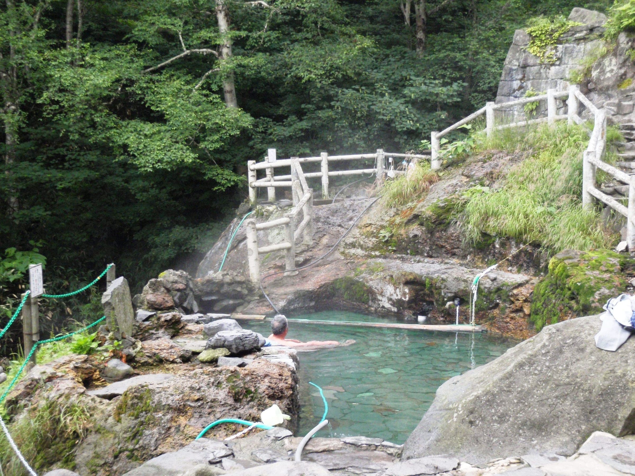 A turquoise rock pool steams below a wooden railing in the woods at Fukiage Roten-no-yu