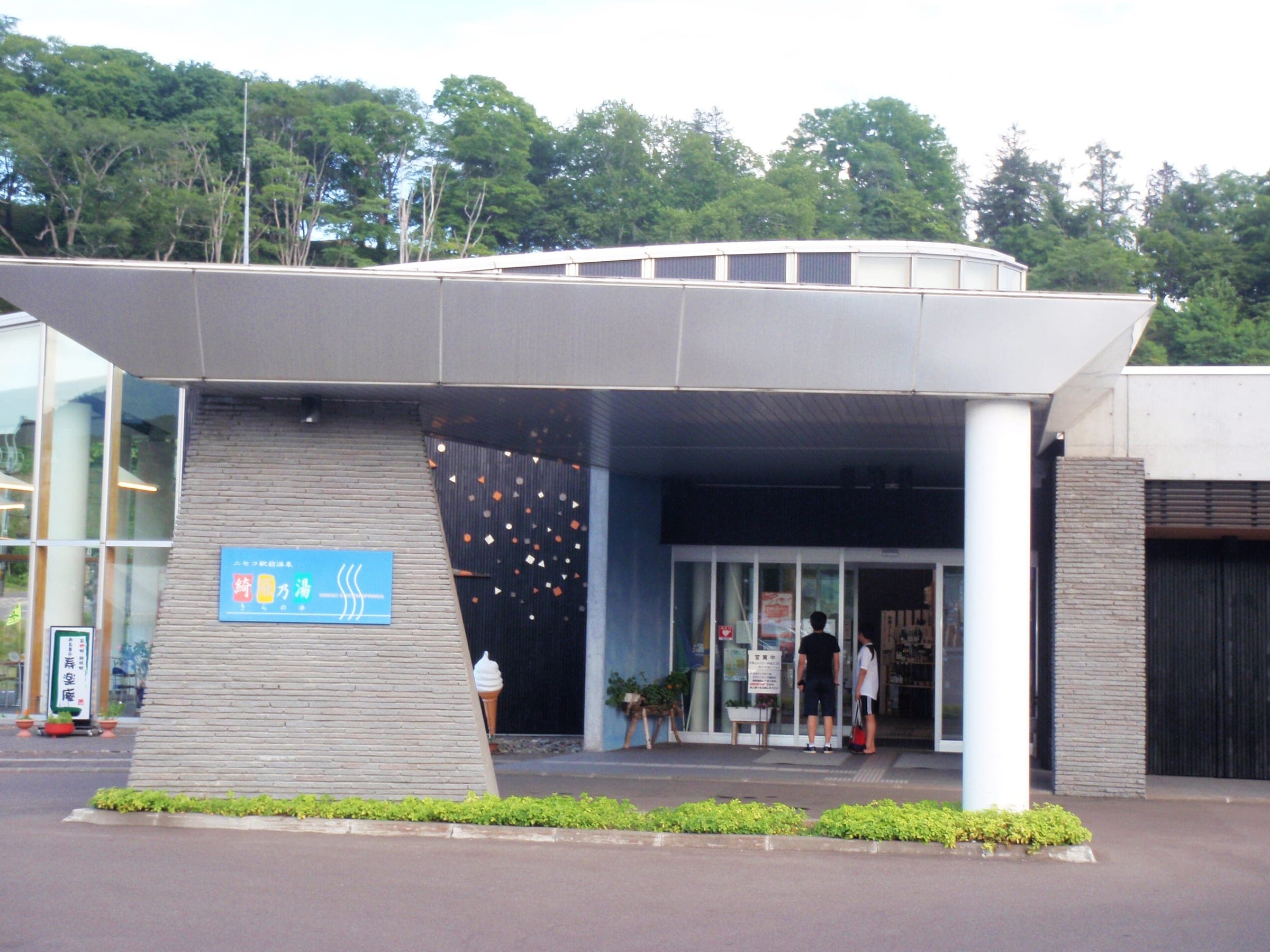 The glass and concrete entrance of Niseko Ekimae Onsen Kiranoyu on a summer day