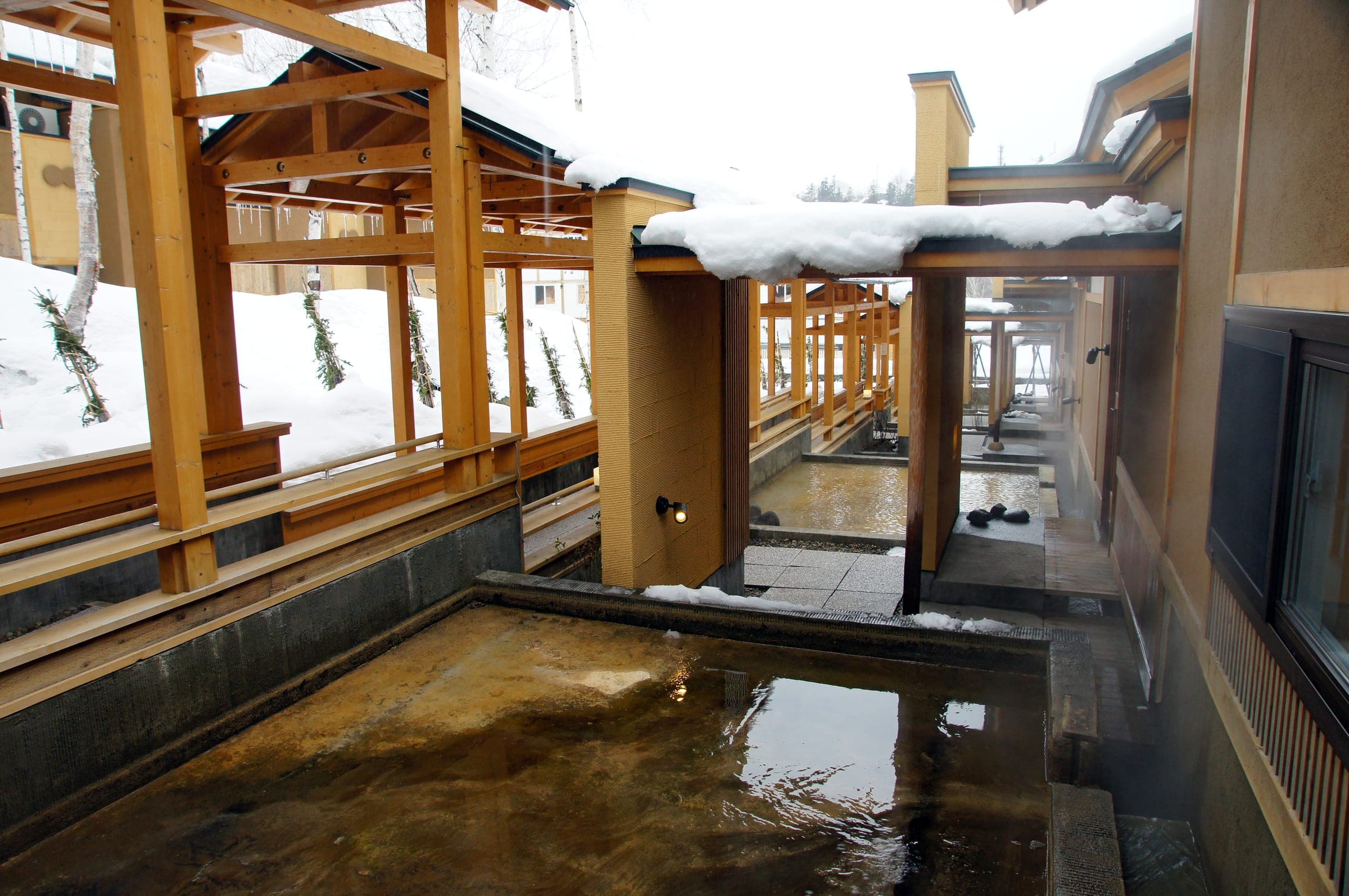 A timber walkway runs beside a shallow bath under snowy eaves at Shirogane Onsen