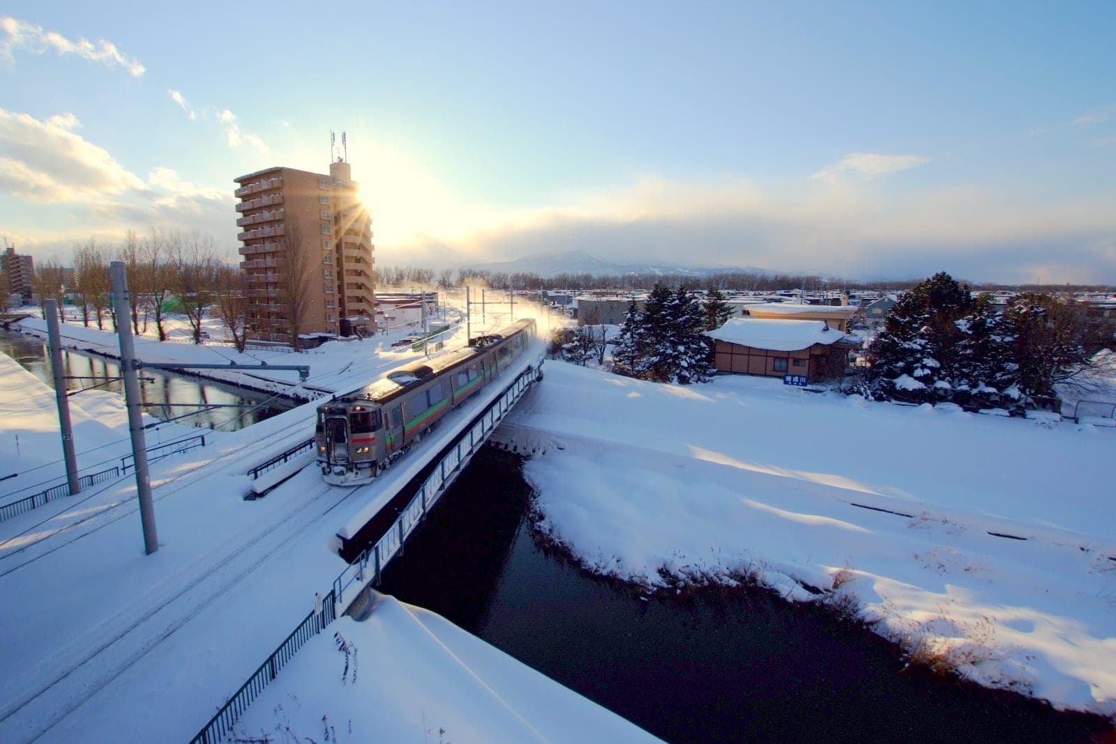 A JR Hokkaido Gakuen Toshi Line train crosses a snowy river near Sapporo in low winter sun
