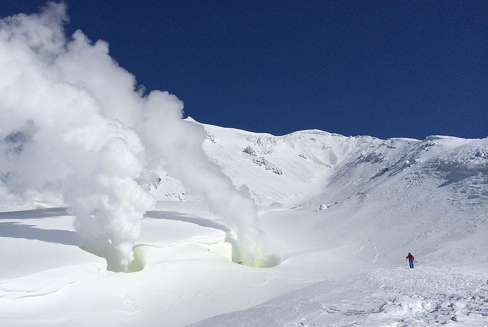 Fumaroles steam against a deep blue sky as a lone skier crosses the snowfield on Asahidake