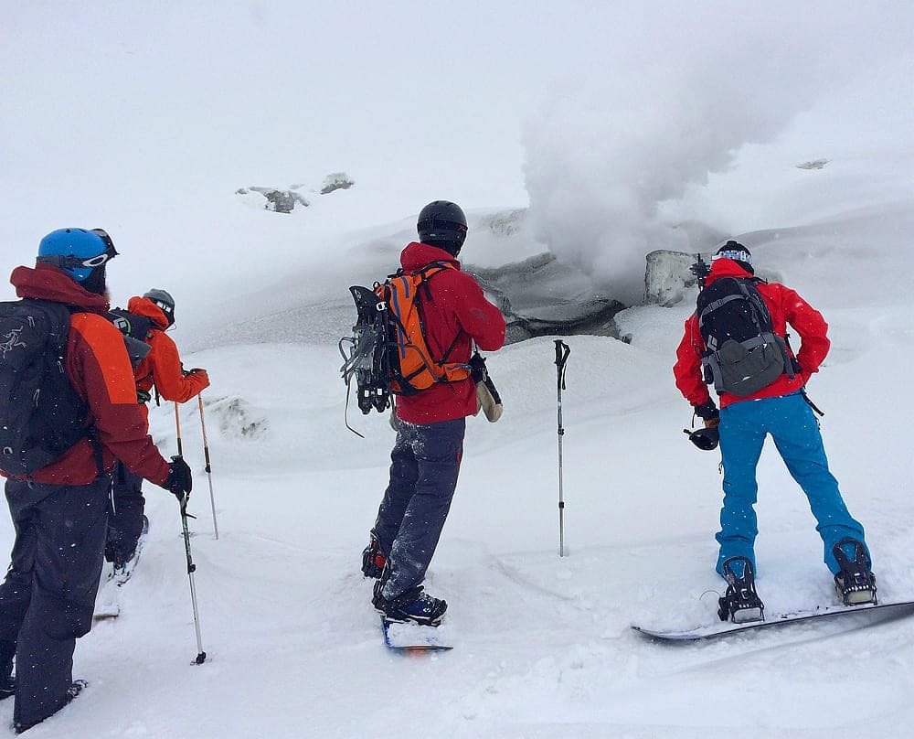 A snowboarder and skiers stand beside a steaming vent high on Asahidake in falling snow