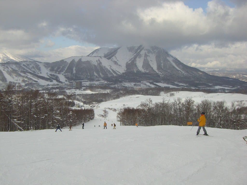 Skiers ride a broad run at Rusutsu below a ridged snowy peak under heavy cloud