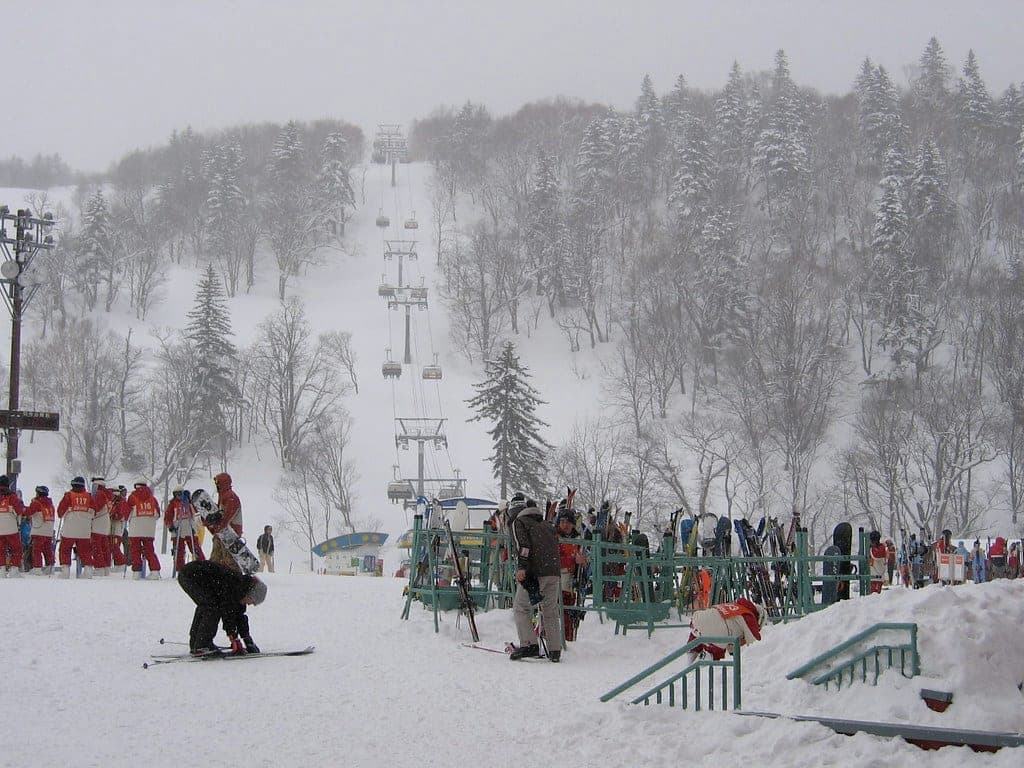Skiers queue at the lifts below snow-covered trees on a grey day at Sapporo Kokusai