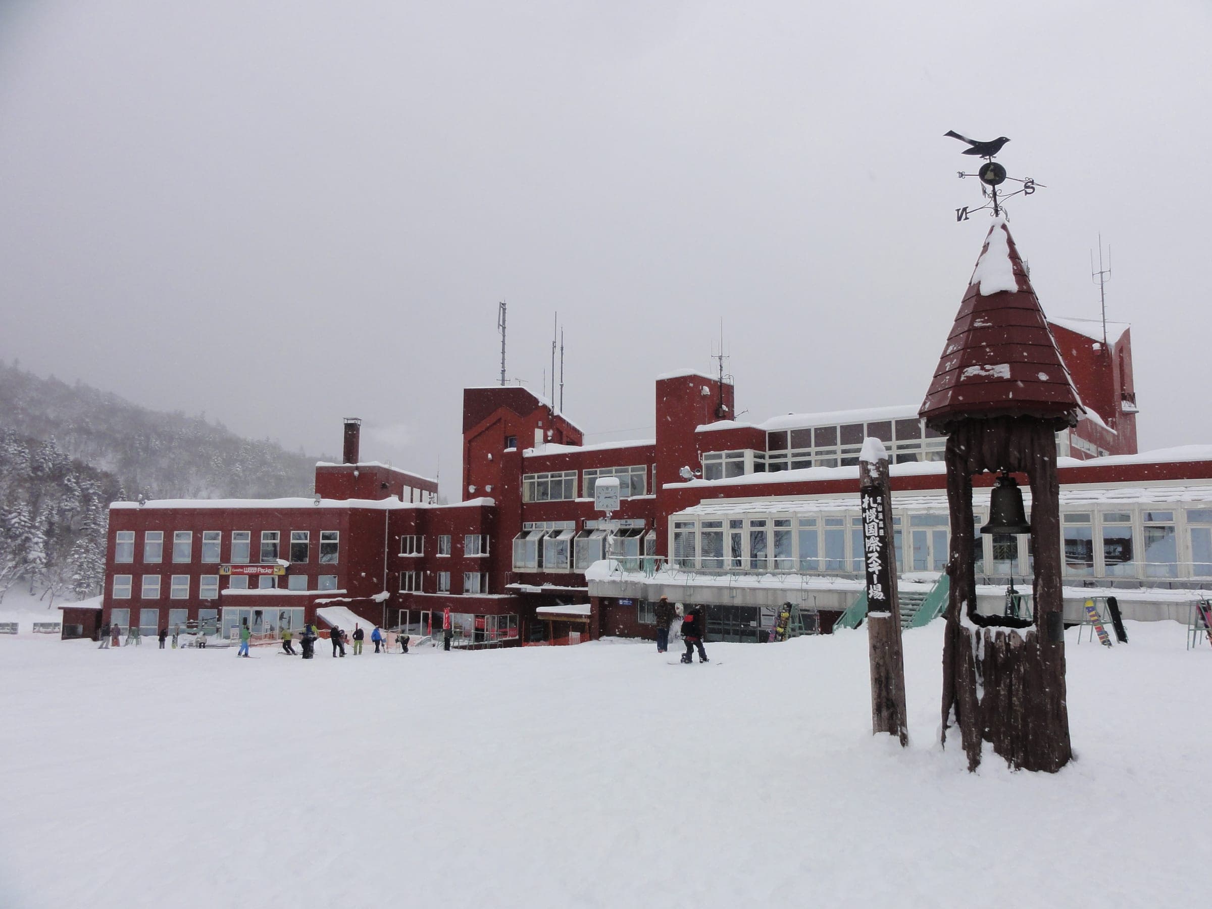 The red-roofed base lodge at Sapporo Kokusai stands in falling snow beside a clock tower