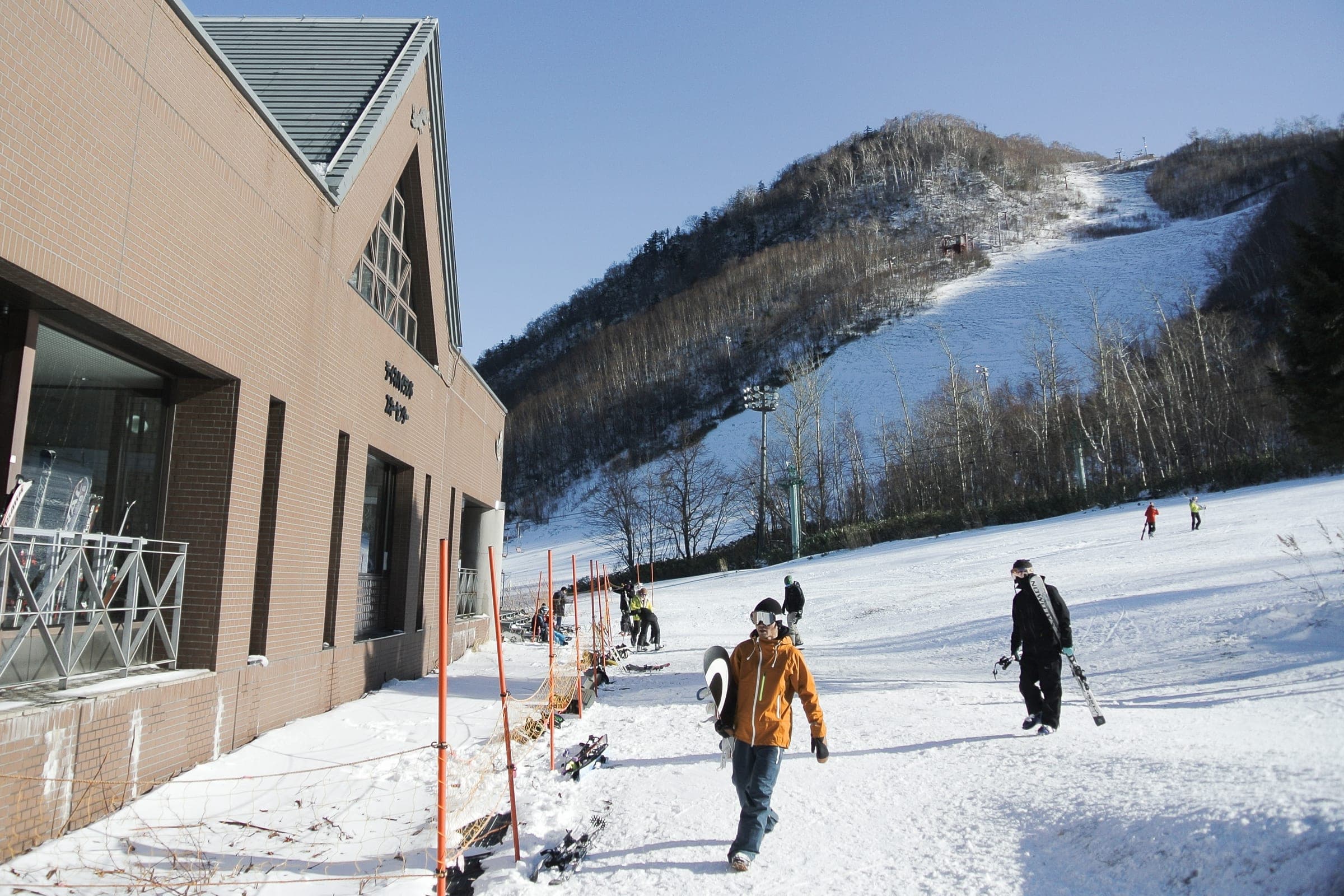 Snowboarders walk past the base lodge at Sapporo Teine below a steep groomed run