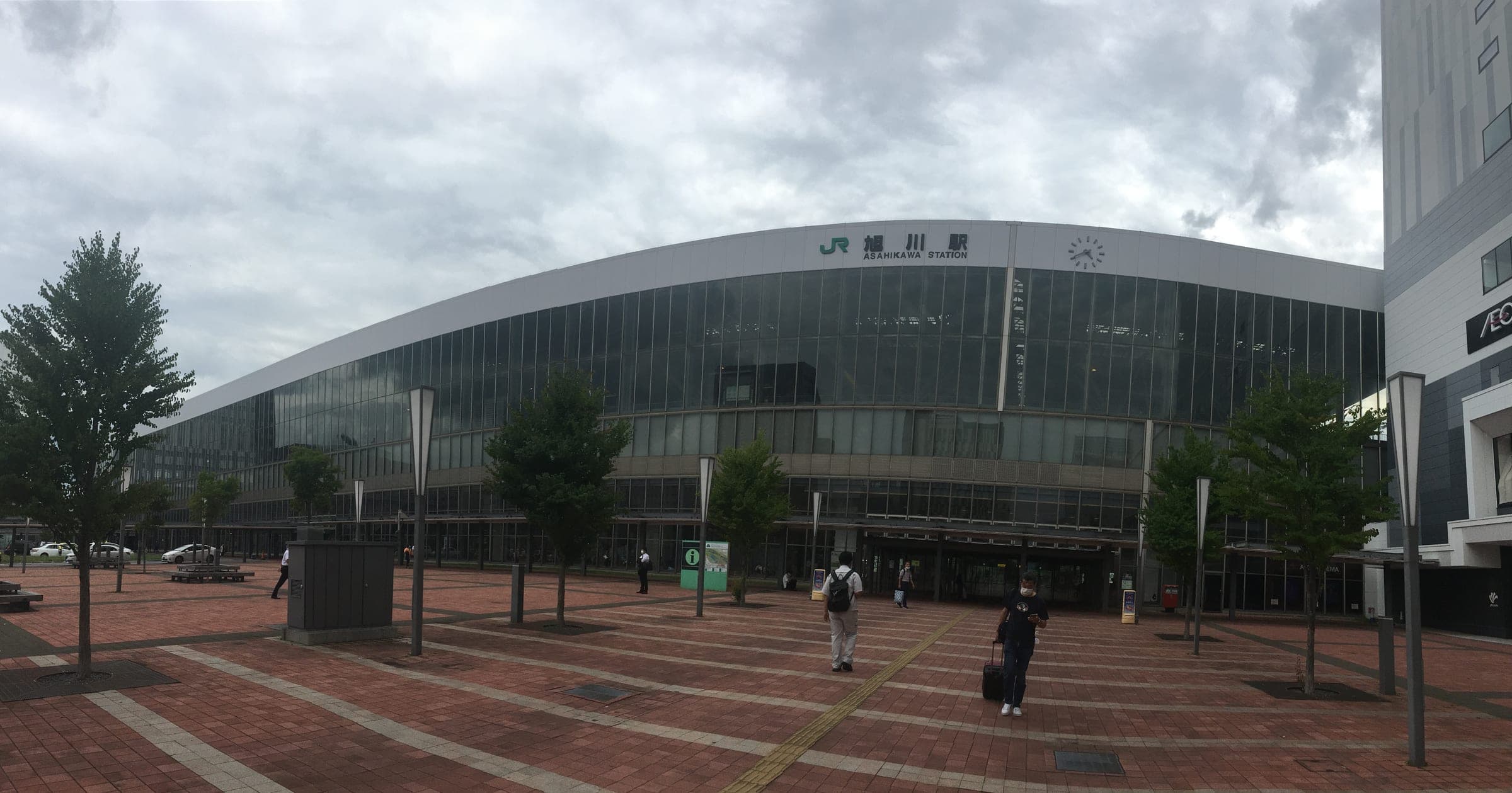 The curved glass facade of Asahikawa Station across its brick plaza in summer