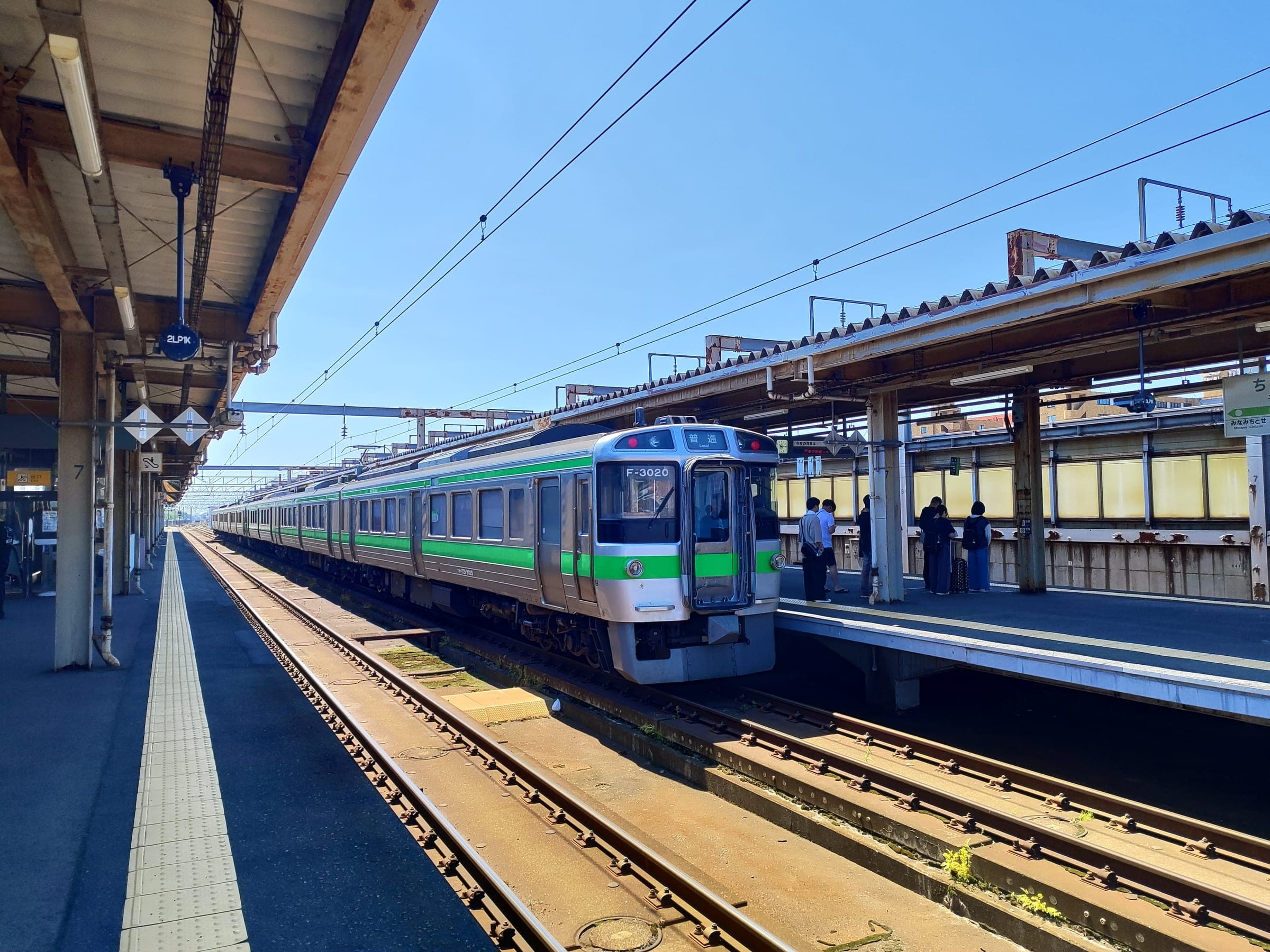 A green and white local train waits at the elevated platform of Chitose Station in summer