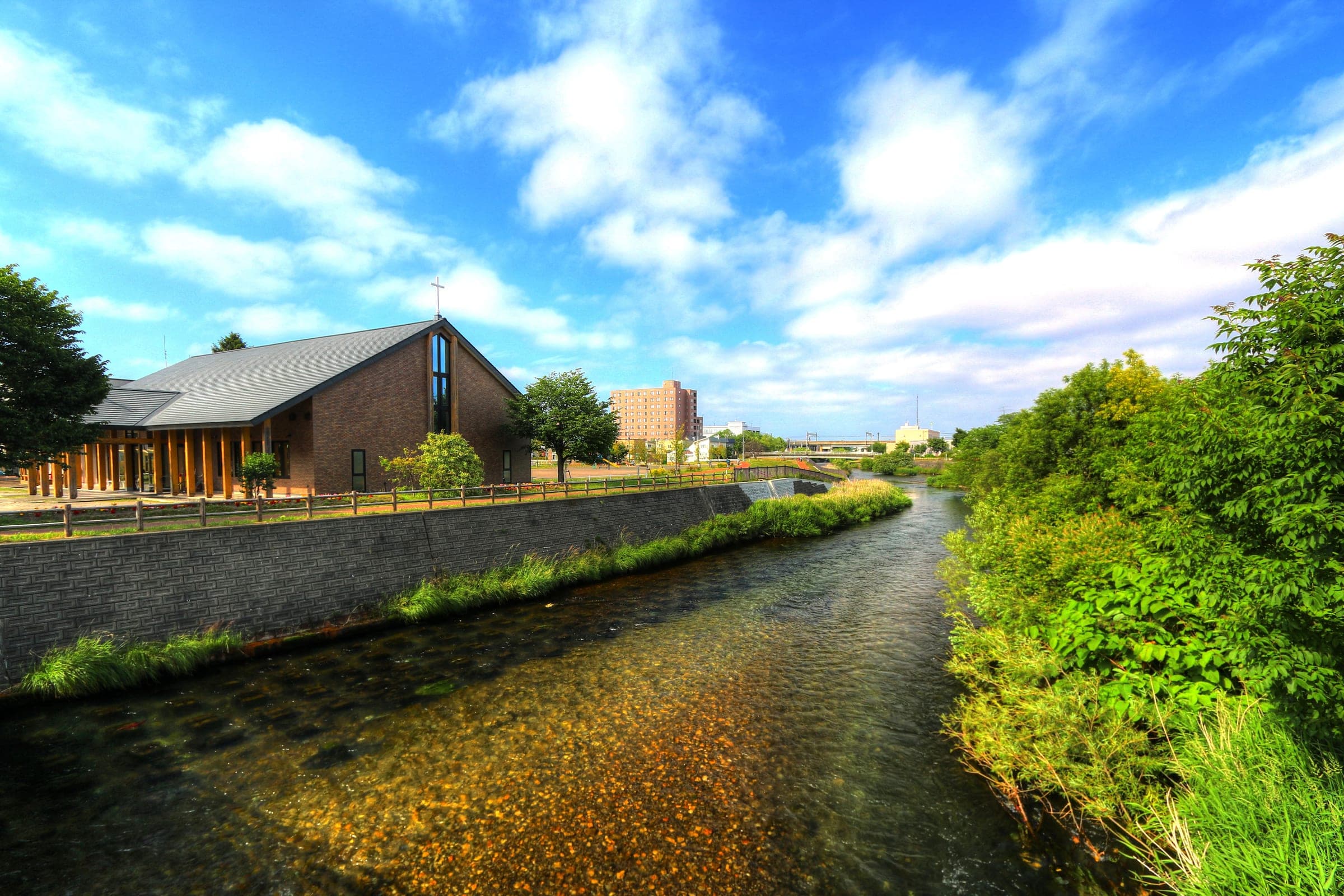 The Chitose River runs between grassy banks past a timber church in central Chitose on a summer day