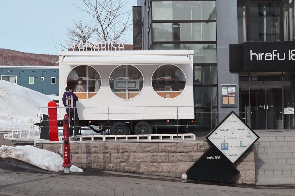 The white % Arabica kiosk with round windows beside the Hirafu 188 building in snow