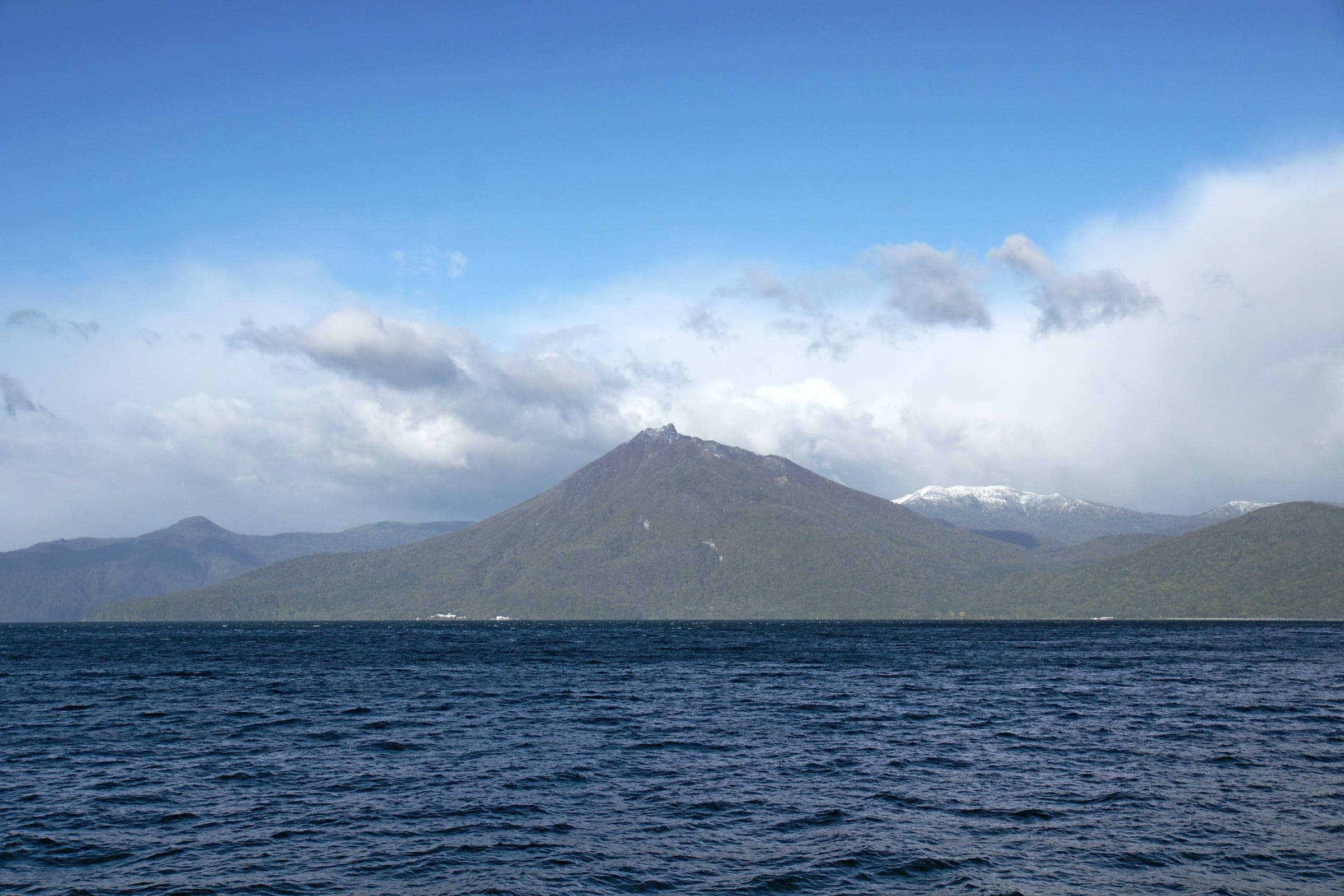 Mt. Eniwa rises beyond the deep blue water of Lake Shikotsu under broken cloud