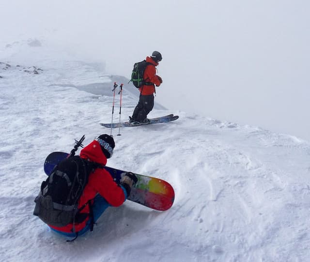 A snowboarder straps in beside a skier on the wind-scoured upper slopes of Asahidake