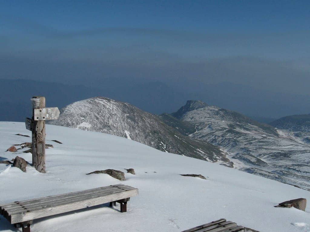 A signpost and bench stand in fresh snow at Hokuchin-dake, looking east toward Kurodake