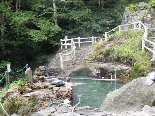 A turquoise rock pool steams below a wooden railing in the woods at Fukiage Roten-no-yu