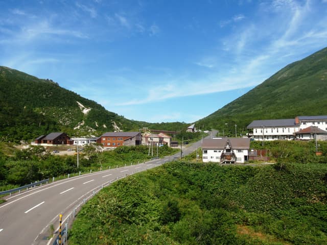 The road climbs past Niseko Goshiki Onsen Ryokan and its neighbouring lodge between green summer slopes