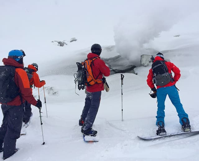 A snowboarder and skiers stand beside a steaming vent high on Asahidake in falling snow