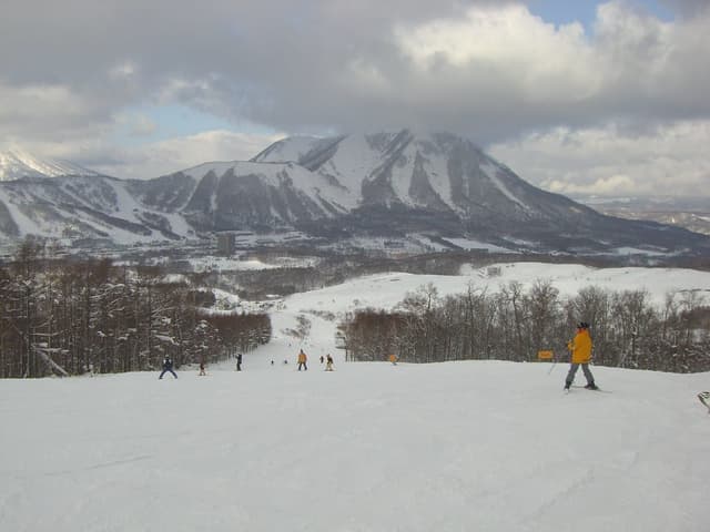 Skiers ride a broad run at Rusutsu below a ridged snowy peak under heavy cloud