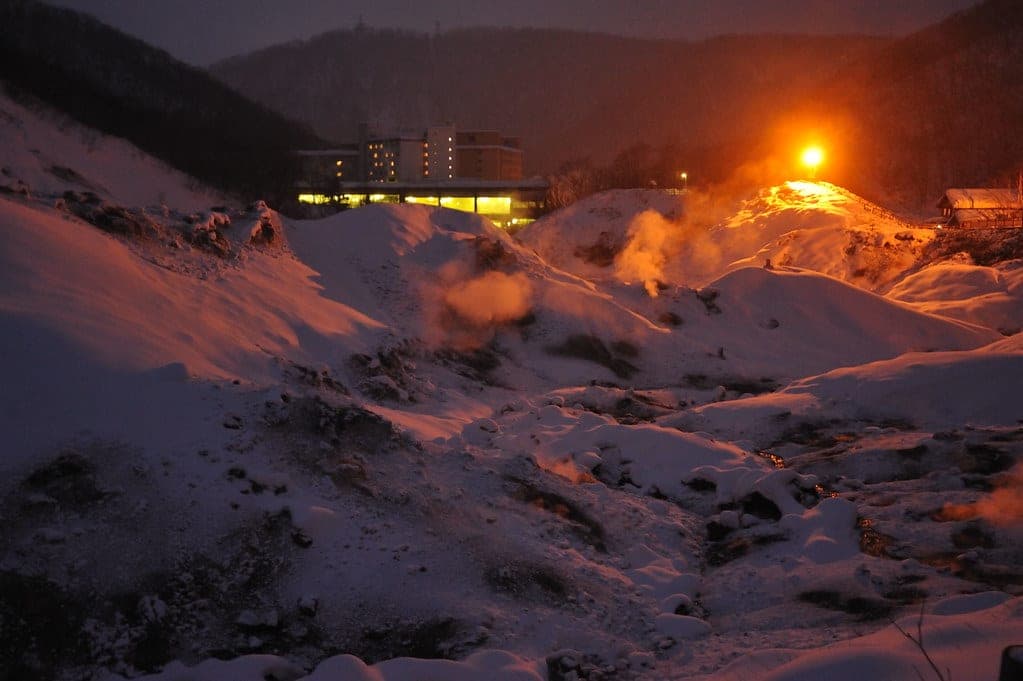 Lights from the Noboribetsu Onsen hotels glow over snow and steam in Jigokudani at dusk