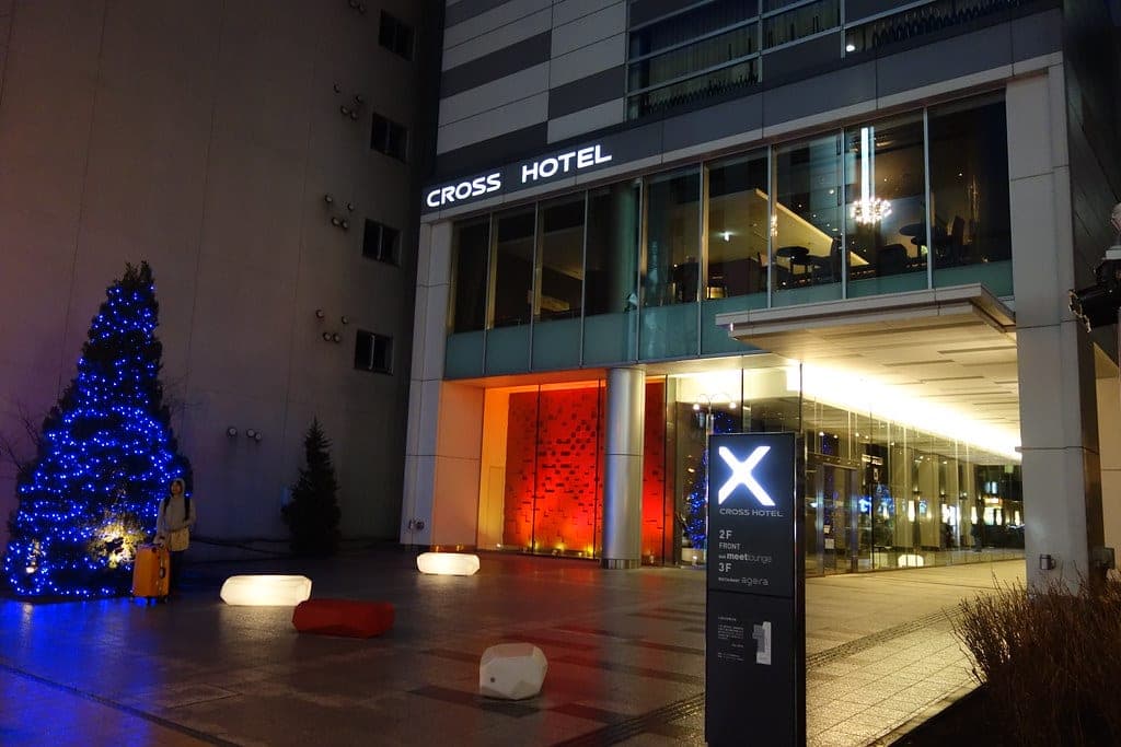 Cross Hotel Sapporo's X sign and glass entrance glow at night beside a blue Christmas tree on the forecourt