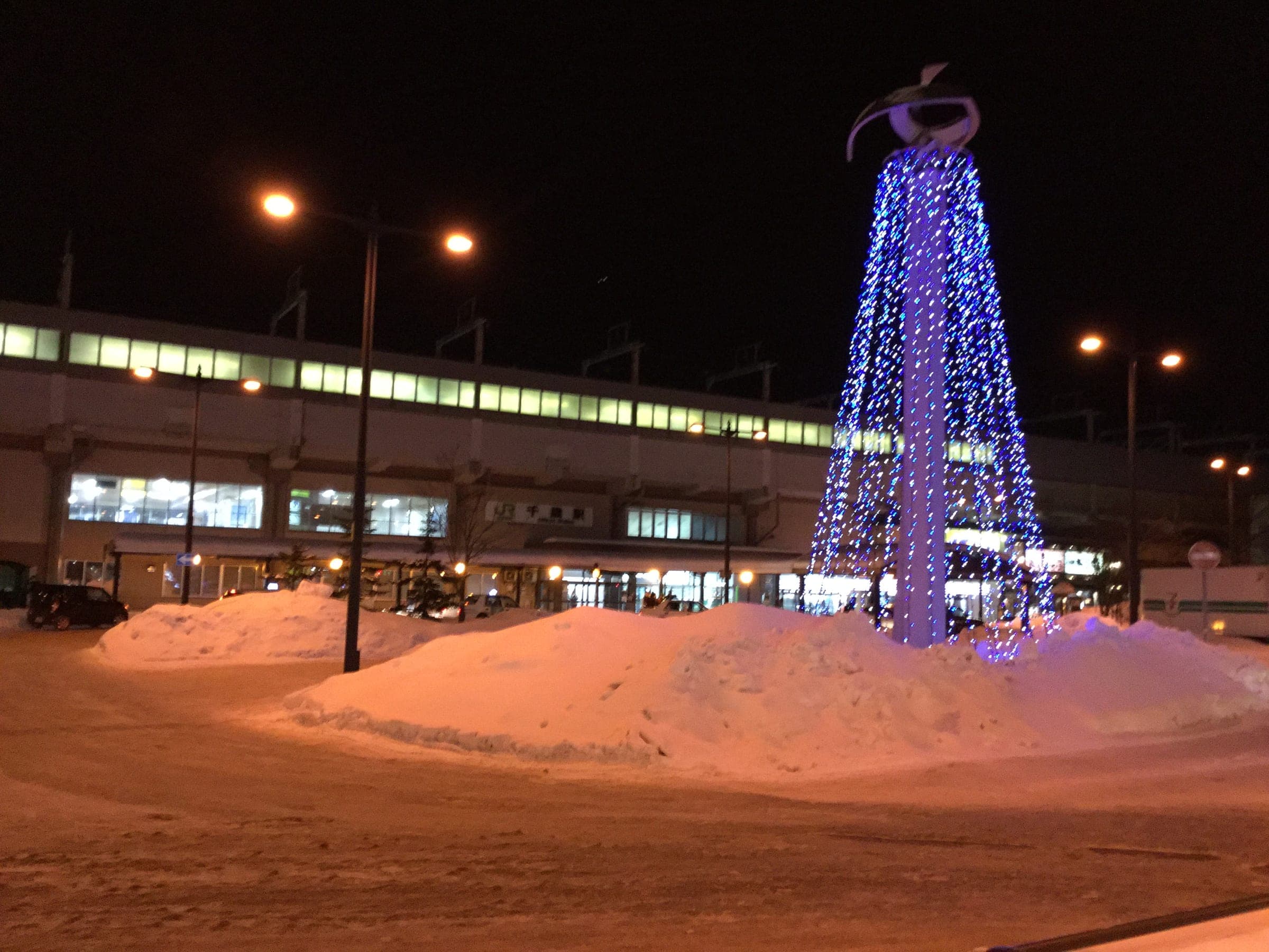 A lit Christmas tree rises from banks of snow outside Chitose Station on a winter night