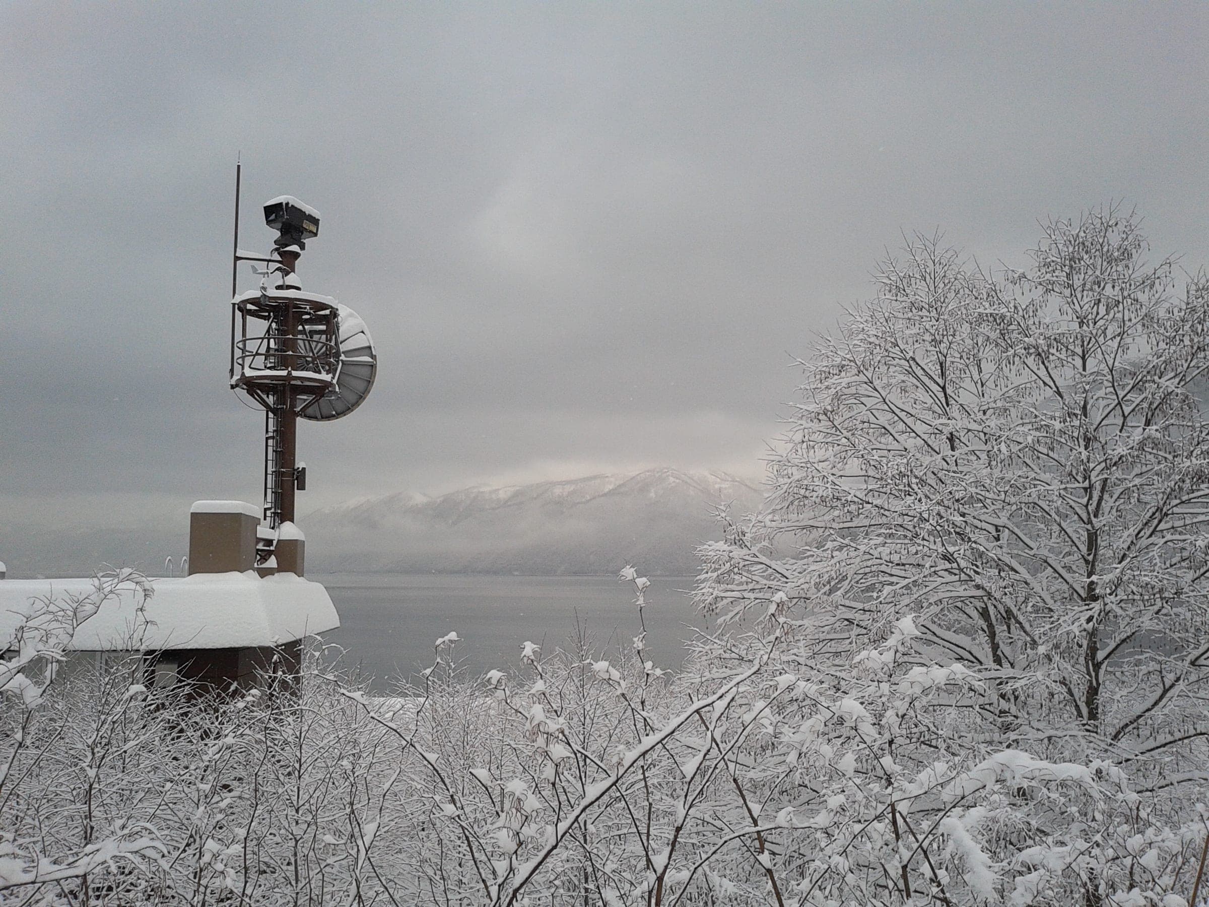 Snow weighs down lakeside trees above the grey water of Lake Shikotsu in winter