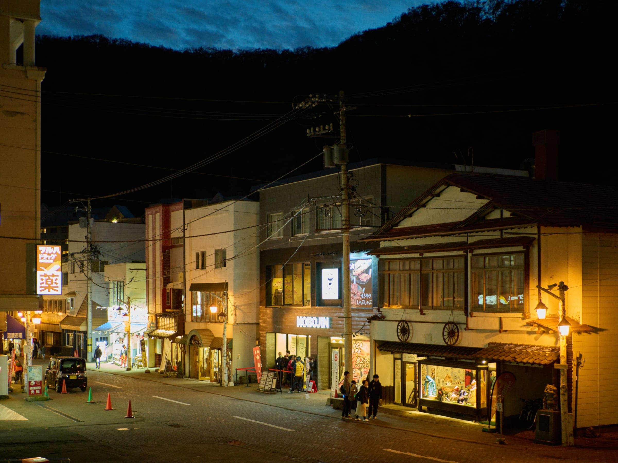 Lit shopfronts including NOBOJIN line the main street of Noboribetsu Onsen at dusk