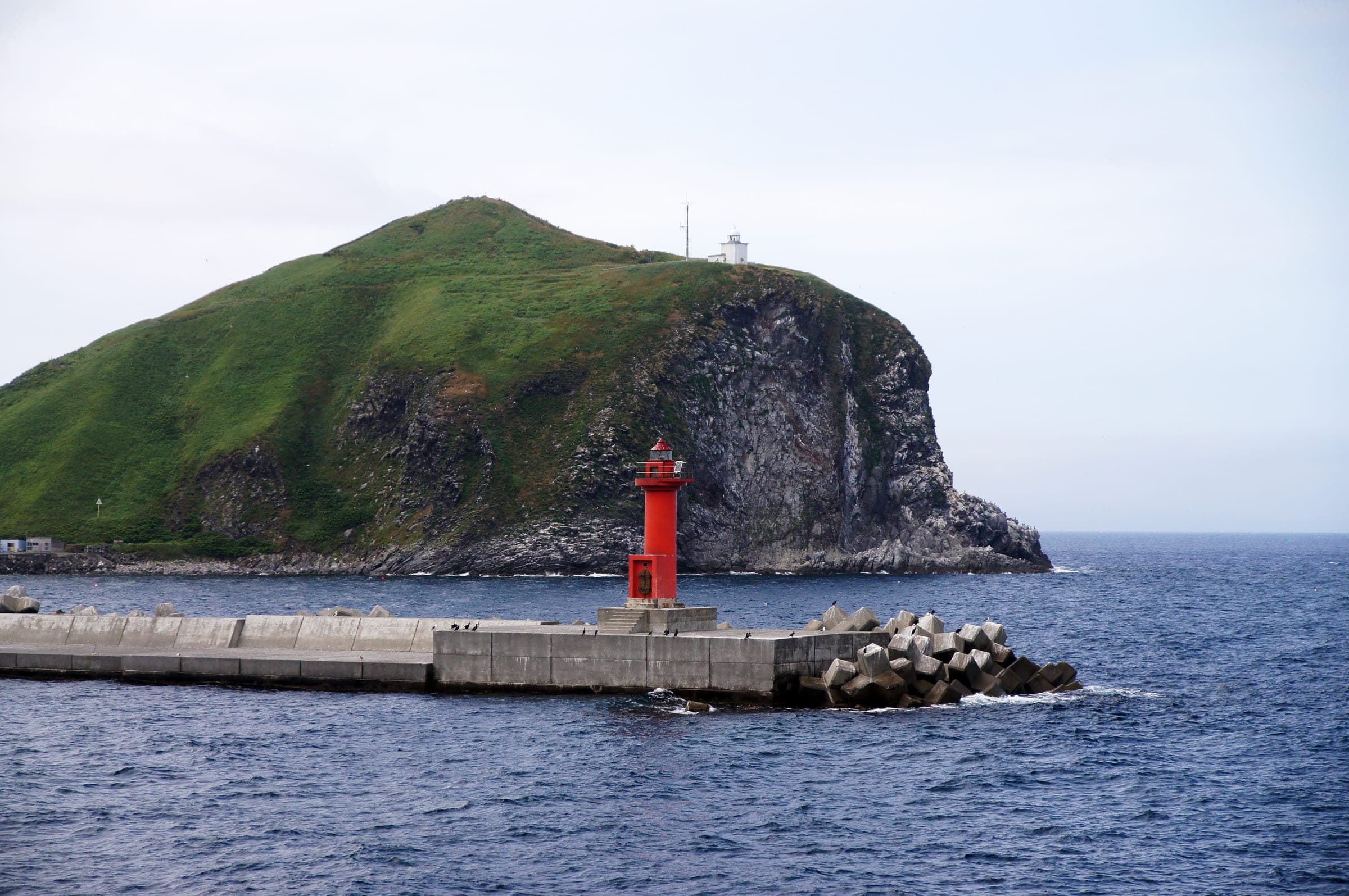 A red lighthouse at the harbour mouth below the green bluff of Cape Peshi on Rishiri